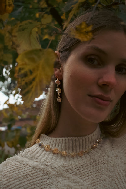 Woman wearing a beige sweater and gold earrings with leaves in the background