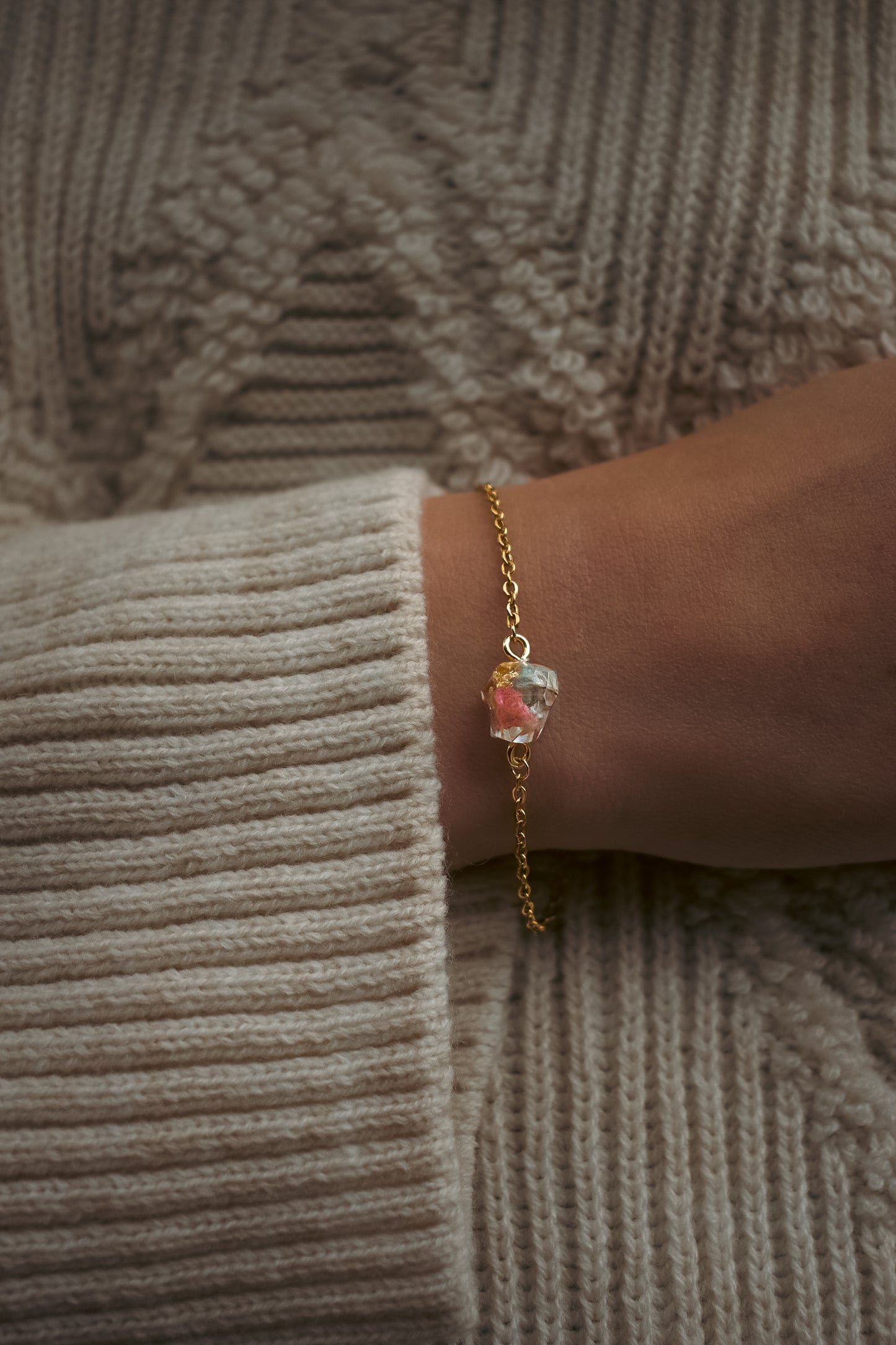 Gold bracelet with a colorful gemstone worn on a wrist against a textured beige sweater.