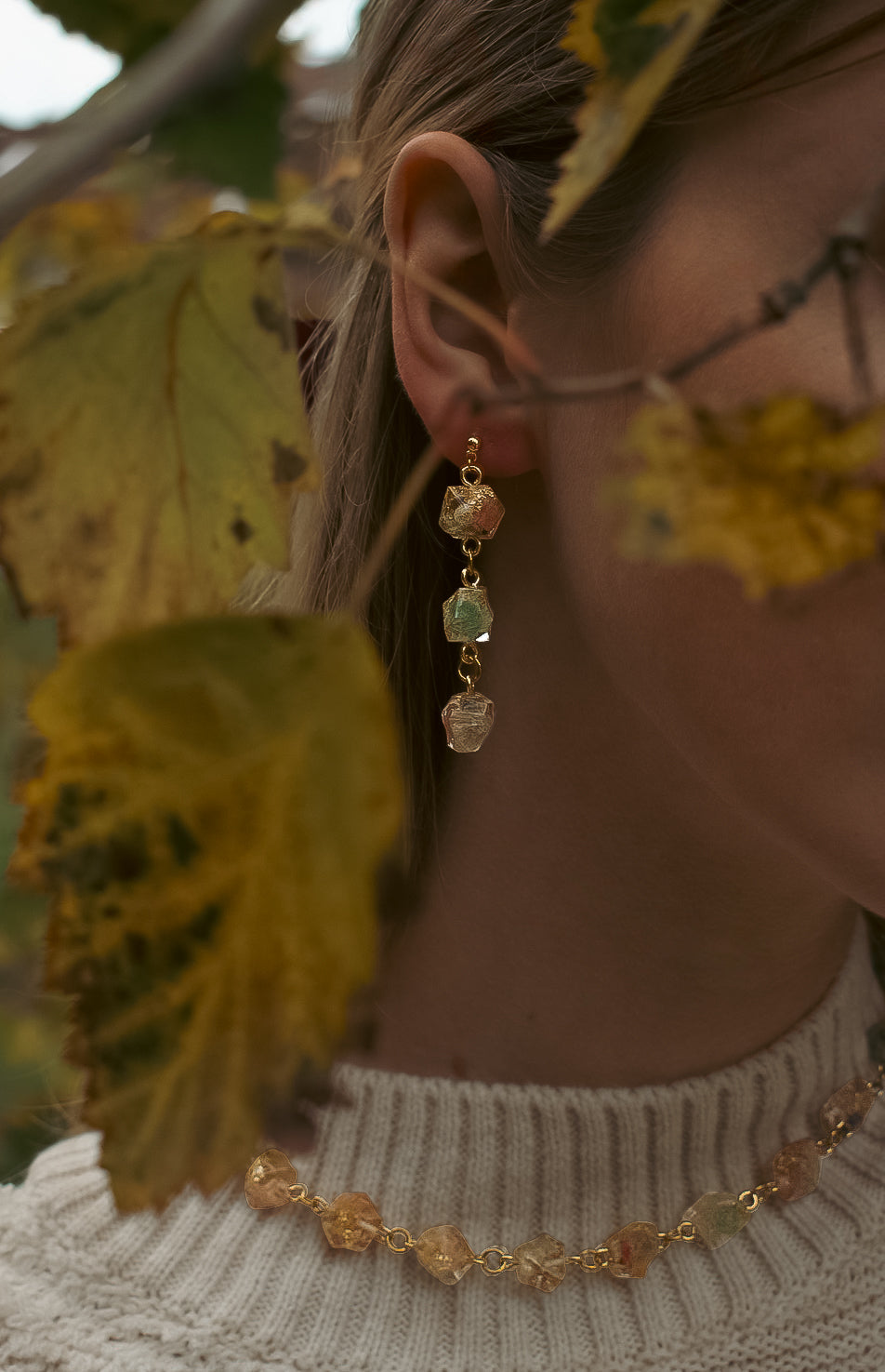 Close-up of a person wearing gold earrings with gemstones against a blurred natural background.