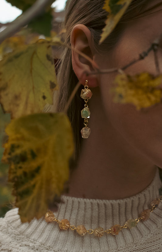 Close-up of a person wearing gold earrings with gemstones against a blurred natural background.