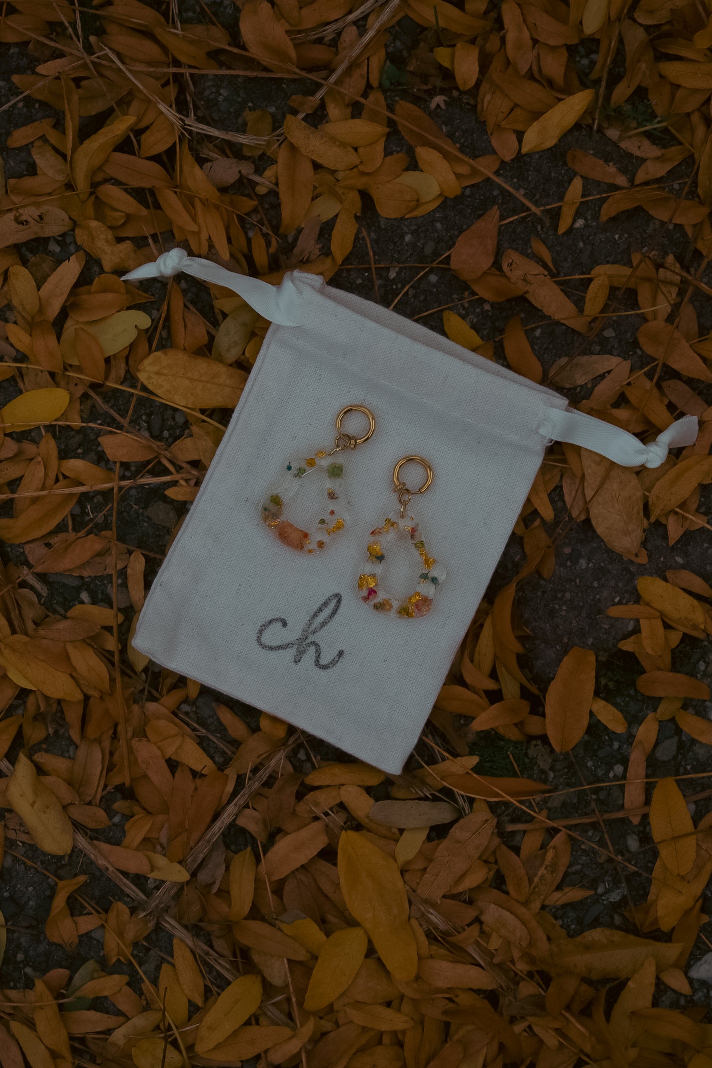 Pair of earrings on a white drawstring bag with 'ch' on it, lying on a ground covered with orange leaves.