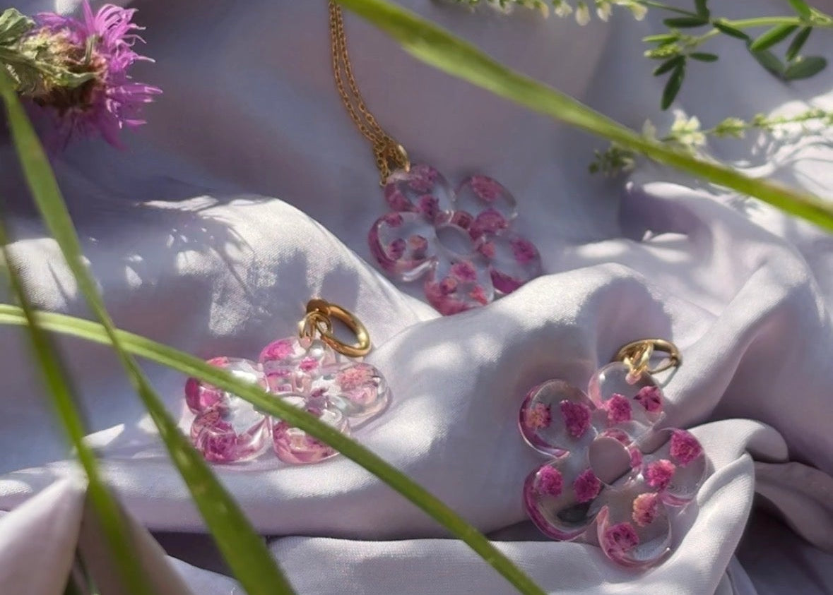 A gold necklace and matching earrings with pink floral heart-shaped pendants, styled on draped white fabric with green grass and wildflowers in the background.