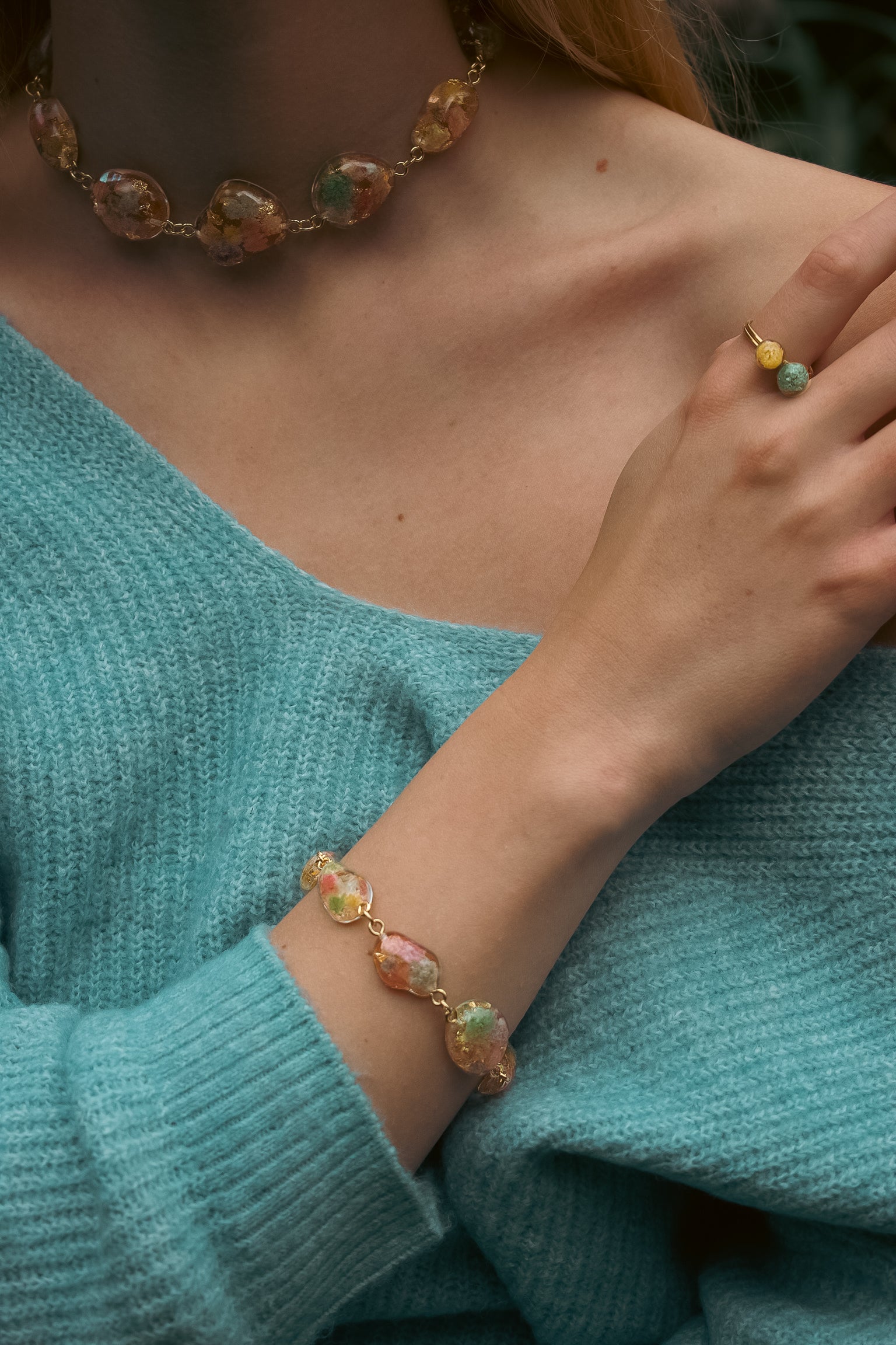 Close-up of a person wearing a colorful beaded bracelet, necklace, and ring against a teal sweater.