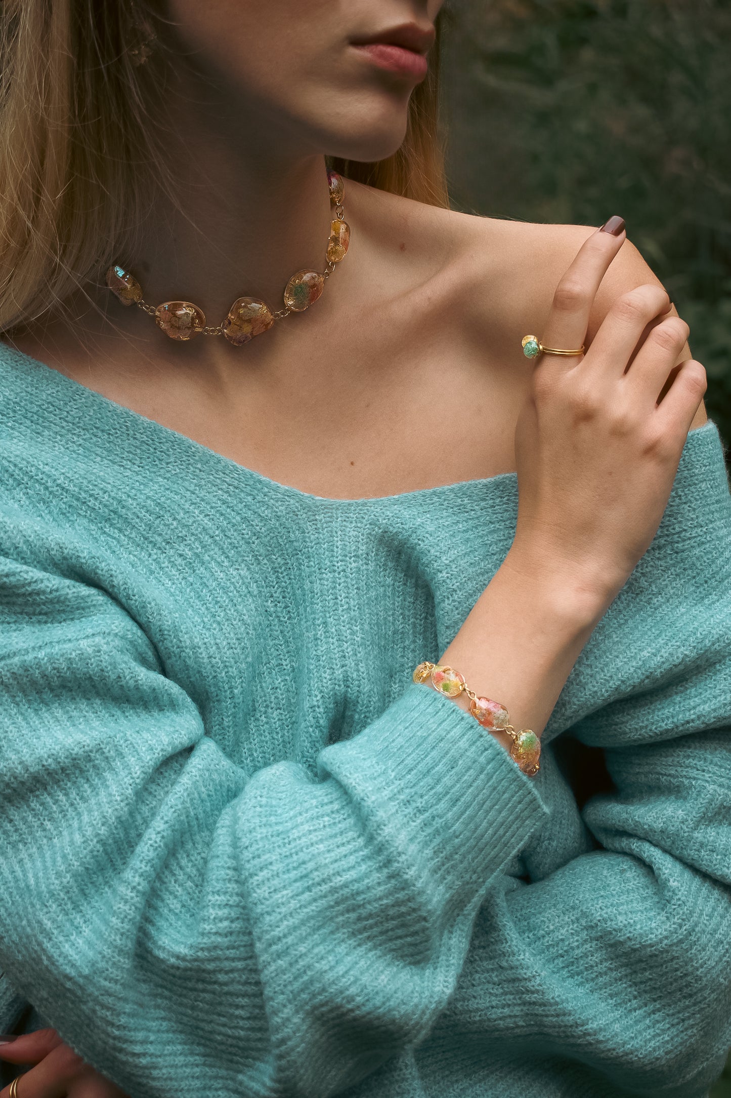 Close-up of a person wearing a turquoise sweater with jewelry including a necklace, ring, and bracelet.