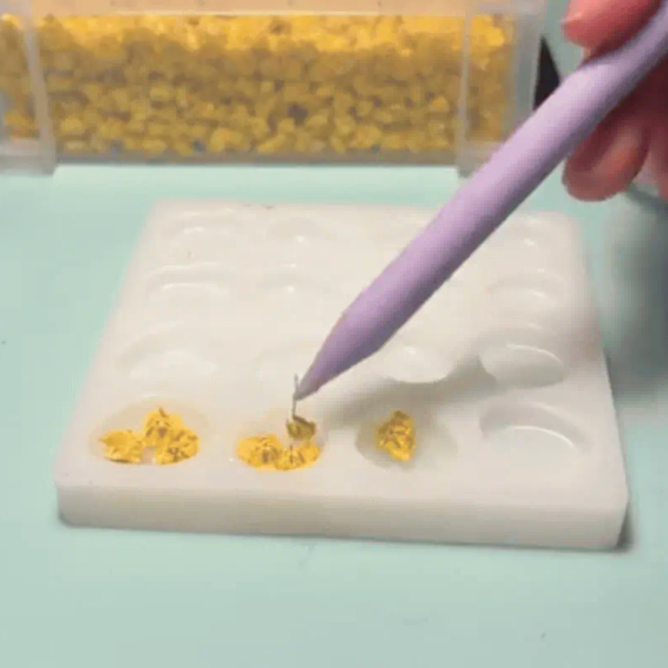 A hand holds a lavender pen-like tool over a white silicone mold tray containing small yellow flowers, with a clear container of more yellow flowers in the background.