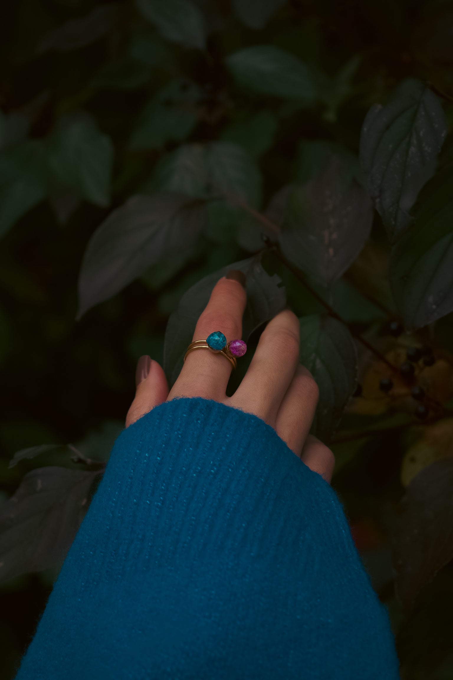 Hand wearing a ring with blue and pink stones against a dark leafy background